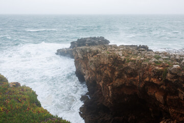 Powerful ocean waves crash against rugged cliffs in Cascais, Portugal, sending sea spray into the air under a cloudy sky. Stock photo