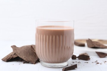 Tasty chocolate milk in glass and pieces of chocolate on white tiled table, closeup