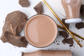 Tasty chocolate milk in glass, bottles and pieces of chocolate on white tiled table, flat lay