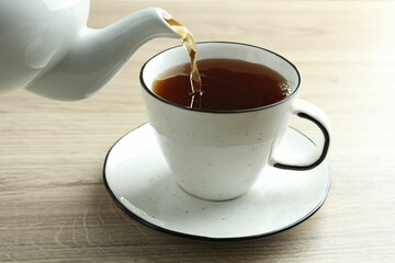 Pouring freshly brewed tea from teapot into cup at wooden table, closeup