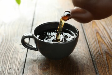 Pouring freshly brewed tea from teapot into cup at wooden table, closeup