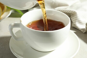 Pouring aromatic tea from teapot into cup at light textured table, closeup