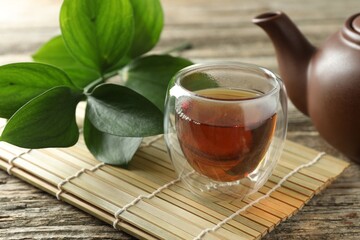 Aromatic tea in cup, bamboo mat, branch with leaves and teapot on wooden table, closeup