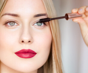 Fototapeta premium Woman applying mascara with confidence, defined brows and red lipstick. Blonde beauty focused on her eye makeup, cosmetics look