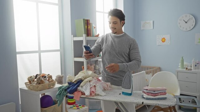 Young man taking selfie in cozy bedroom surrounded by baby items, an ironing board, and a crib, capturing a moment of daily life in a warm, homey environment.