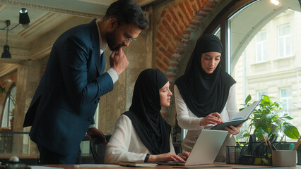 Three coworkers colleagues businessman businesswomen collaborate work together Arabian man muslim women hijab male female teamwork discuss talk write typing laptop business project at office company