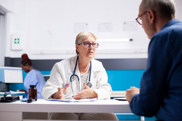 Obraz premium Male pensioner patient sits across from retired lady doctor in clinic discussing treatment plans. Senior female physician sitting at desk and giving medical advice to old white man in hospital room.