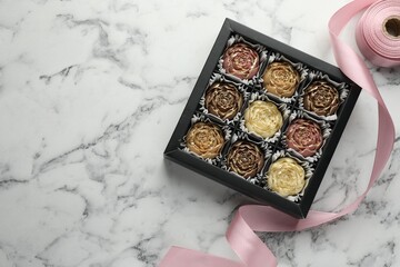 Delicious flower shaped chocolate bonbons in box and ribbon on white marble background, top view. Space for text