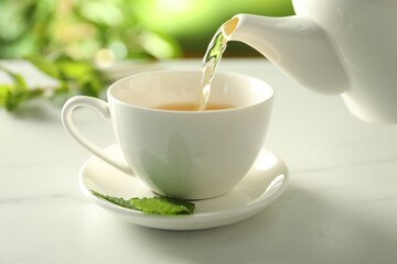 Pouring freshly brewed tea from teapot into cup at white marble table, closeup