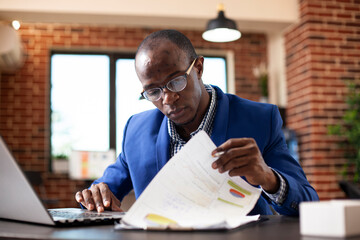Closeup of black man seated at office desk, deeply focused on analyzing financial documents in...