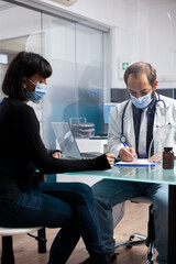 Fototapeta premium Young woman interacts with physician at clinic office, focused on understanding her recovery plan. Female patient observing doctor write on clipboard with protection screen separating them at desk.