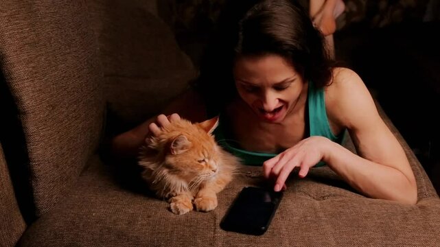 Beautiful woman holds mobile phone for online work, chating , text, talk. Happy fun joying girl lying on sofa at home interioor with her orange fur cat, peting and kiss. Emotional people closeup video