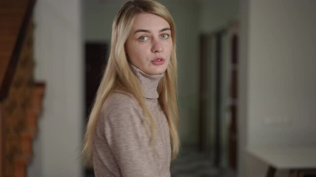 A blonde woman holds a bundle of cash out to the camera with a serious expression. She is indoors, with wooden stairs in the background. Her gesture and expression suggest that she is hinting her