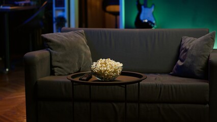 Bowl of popcorn on table next to remote control in neon illuminated apartment home theater. Close...