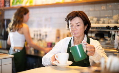 Senior woman cafe worker pours coffee from cezve coffee pot into cup, prepares order for customer. Barista finishes creating hot drink in coffee shop