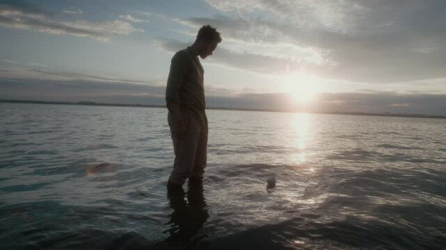 Wide shot with lens flare of young African American guy standing in lake water at sunset, launching origami paper boat, then looking at it sail away on rolling waves, in glaring sun