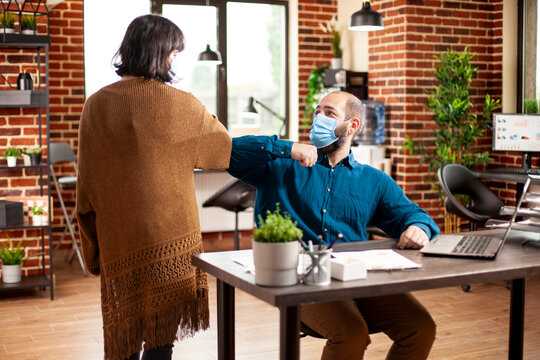 Young man and woman in business casual clothing greet each other with elbow bump at desk in brick wall room. Caucasian colleagues wearing face masks and ensuring pandemic safety in startup office.