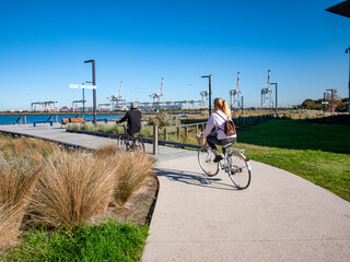 A scenic bicycle path or walking and cycling trail in Port Melbourne, Australia, with two young cyclists riding along a paved footpath and industrial port infrastructure in the distance