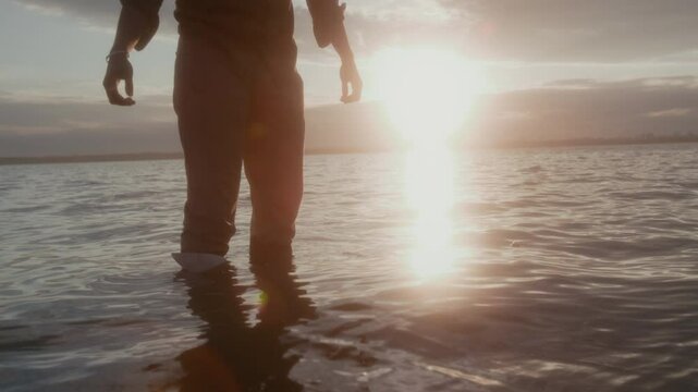 Medium tilting down shot with lens flare of young black man standing in lake on summer day, sailing paper boat and watching it swim away on rocking waves, against bright sun light