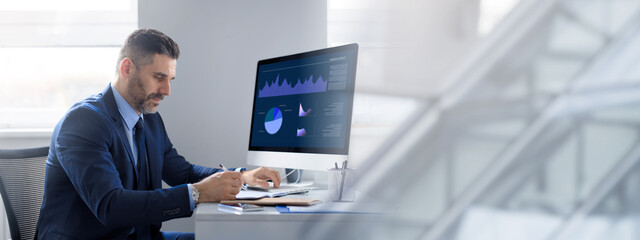 A middle aged businessman focused on his tasks at a well-organized office desk. He is analyzing data on a computer screen while taking notes, showcasing a professional lifestyle, copy space