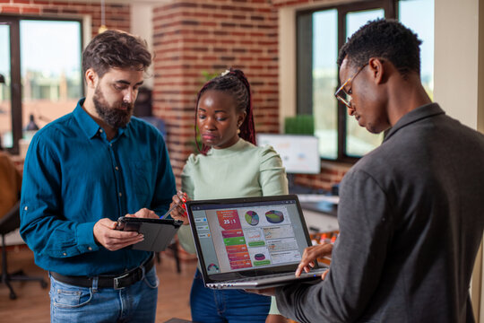 Colleagues utilizing digital technology during their discussion in modern workspace. Black man using laptop to retrieve marketing data as his male and female coworkers review strategy plans on tablet. - Powered by Adobe