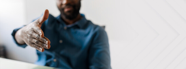 A young African American businessman extends his hand in a gesture of partnership. He is seated at a sleek desk in a contemporary office environment, embodying success and professionalism, copy space