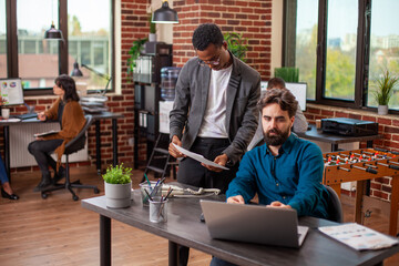 African american businessman with clipboard discusses marketing ideas with male colleague in a brick wall office. Bearded man types on laptop and listens carefully to suggestions from black coworker.