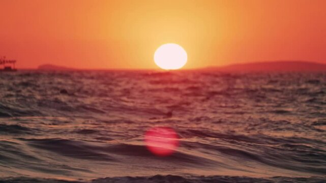 Minimalist tripod shot of the sun setting directly above the ocean horizon, casting a fiery orange glow over distant waves. A dramatic silhouette of the sea is visible in the foreground