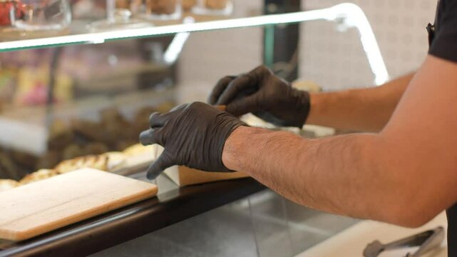 Close up of hands of takeaway shop employee handing a paper food package with cookies to a customer over glass counter. Cozy small business scene focused on artisanal service. From the back, 4k video.