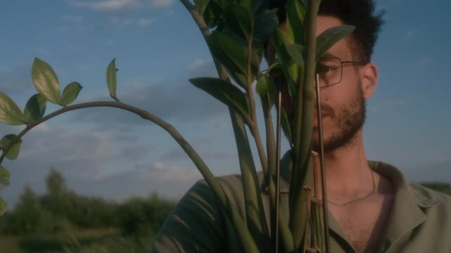Medium closeup portrait of handsome African American man in glasses, with beard standing outdoors in field, holding potted zamioculcas plant and looking at camera through green leaves
