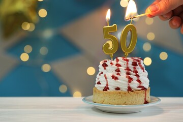 50th birthday. Woman lighting number shaped candles on cupcake at white wooden table against blurred lights, closeup and space for text. Bokeh effect