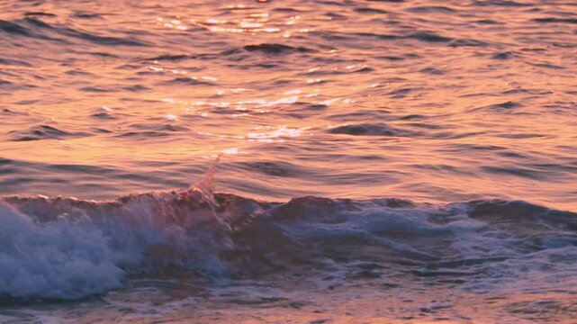 Close-up tripod shot of soft ocean waves reflecting the warm glow of a sunset. The water shimmers with pink and orange tones, creating a calm and abstract seascape perfect for backgrounds