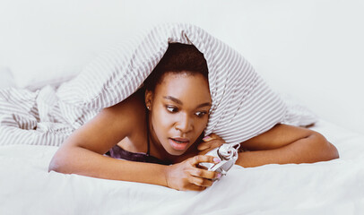 Oversleep concept. Shocked african american girl wakes up, climbs out from under covers looking at alarm clock