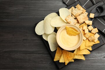Tasty cheese dipping sauce in bowl and different snacks on dark wooden table, top view. Space for...