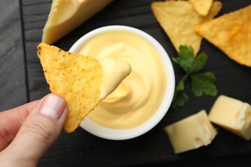 Woman dipping nacho chip into tasty cheese sauce at table, top view
