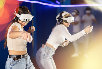 Girls in virtual reality helmets and with joysticks in their hands are playing a video game against guys. Two young women in VR glasses actively playing a new game in the neon light