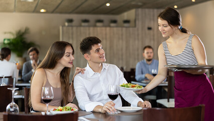 Young woman waitress brings order from couple young man and young woman in restaurant