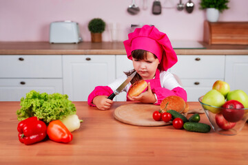 Funny little beautiful child girl in a chef costume prepares burgers while sitting at a table in the kitchen. Healthy eating. Child is cutting sandwich buns with a knife.	