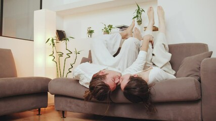 The loving couple playfully lay on the floor near the sofa, legs intertwined, enjoying a tender moment in their cozy living room in the rays of the midday sun.