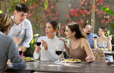 Young man waiter takes order from couple of women and adult man on restaurant terrace