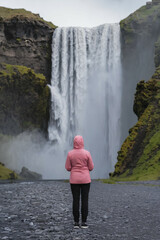 Standing in awe before Seljalandsfoss waterfall, a traveler embraces the tranquil atmosphere. The surrounding lush greenery and cascading water create a magical ambiance on a cloudy day.