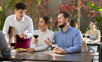 Young man waiter takes order from couple of young man and woman and adult man on restaurant terrace
