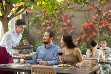 Young guy waiter brings order from couple young man and young woman on restaurant terrace