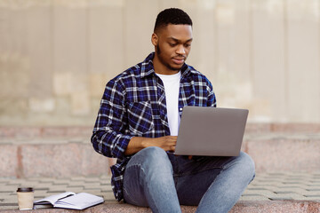Front view portrait of african student using computer outdoor, sitting on steps with coffee cup and planner