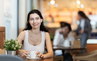 Woman is happy to drink a cup of hot coffee from a white mug in a coffee shop. European woman enjoying cappuccino and sitting at a table in a cafe