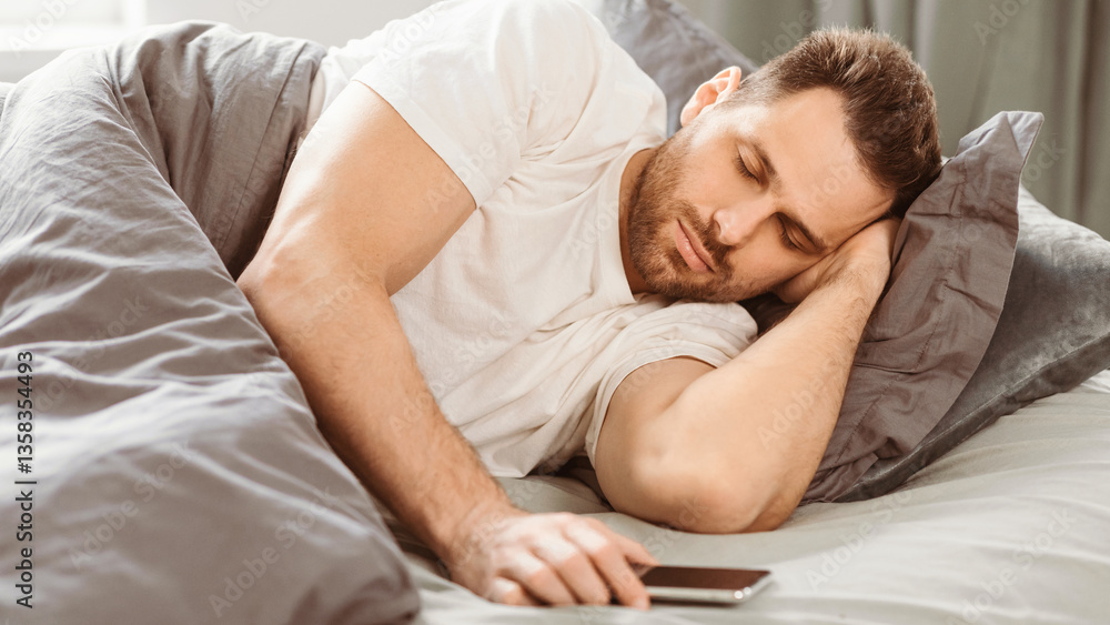 Poster man sleeping througn alarm clock on phone lying in bed at home. smartphone user holding cellphone wh
