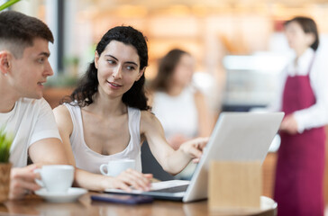 Young man and a woman are sitting at a coffee shop table and discussing the details of a project while looking at a laptop. Man with a woman at a business meeting over a cup of hot coffee.