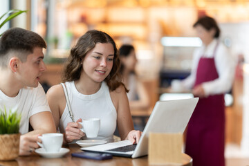 Fototapeta premium Young man and a woman are sitting at a coffee shop table and discussing the details of a project while looking at a laptop. Man with a woman at a business meeting over a cup of hot coffee.
