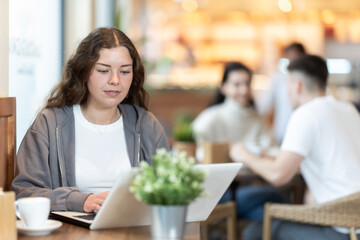 Young woman cafe visitor sits at table with laptop, reads incoming emails and drinks strong coffee. Client is preparing for important meeting, reading text content of report