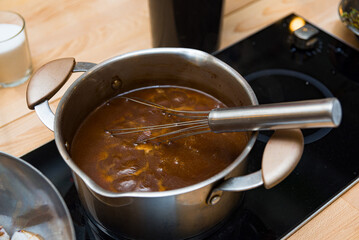Whisk stirring sauce in stainless steel pot. Close-up of a stainless steel pot on a stovetop with a whisk stirring a rich brown sauce, simmering on low heat.